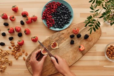 Overhead shot of fresh fruits, nuts, and hands preparing ingredients on a wooden board.