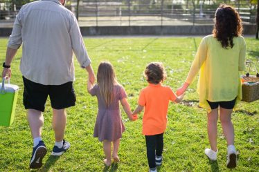 A family of four walking hand in hand through a sunny park carrying picnic supplies.