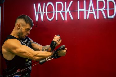 A muscular man exercising in the gym with a motivational 'Work Hard' neon sign on a red background.